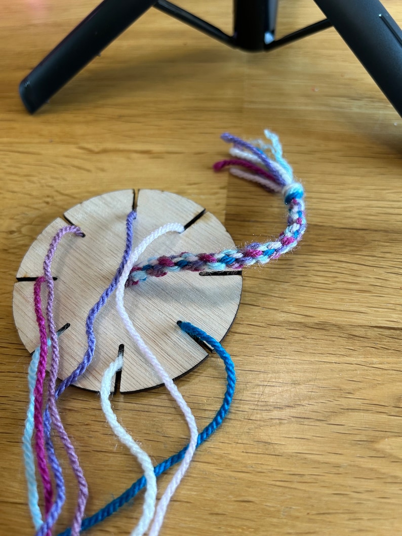 Op de afbeelding: Een houten weefgetouw met zes gleuven voor touw, gebruikt voor het maken van vriendschapsarmbanden. Het weefgetouw heeft vijf touwen die erdoorheen zijn geregen, in de kleuren roze, paars, wit en blauw. Een gevlochten armband wordt gemaakt met de touwen.