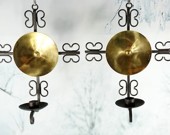 Peut inclure: Deux appliques murales assorties de style vintage. Chacune présente un disque en laiton au centre, entouré de bras en fer forgé en forme de croix avec des accents décoratifs en forme de papillon. Un bougeoir est suspendu en dessous.