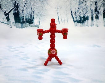 May include: A red wooden candle holder with three arms, each holding a small cup. The candle holder has a decorative floral design in white and green. The base has three legs. The background is a snowy winter scene.