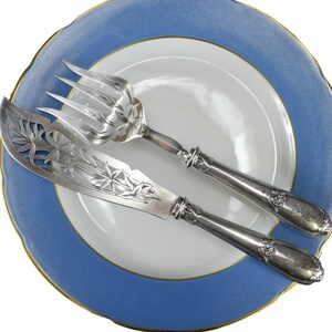 Puede incluir: Un juego de cuchillo y tenedor para pastel de plata sobre un plato azul y blanco. El cuchillo tiene un diseño adornado y el tenedor tiene tres dientes.