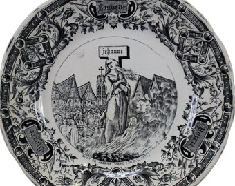 Może przedstawiać: Biały talerz ceramiczny z czarno-białym wzorem. Talerz przedstawia scenę religijną z kobietą trzymającą krzyż i kościołem w tle. Na talerzu widoczny jest tekst "Bonnevie" i "Jeanne".