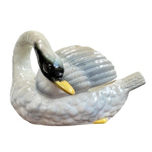 May include: A white porcelain swan figurine with a black head and yellow beak. The swan is curled up with its head tucked under its wing.