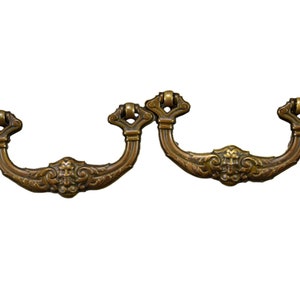 Könnte beinhalten: Zwei antike Messing-Schubladengriffe mit verzierten Designs. Die Griffe haben eine gebogene Form und ein dekoratives Mittelstück.