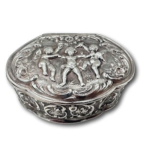 Pode incluir: Uma caixa de joias oval de prata com um design ornamentado que apresenta três querubines de mãos dadas. A caixa é decorada com motivos florais e tem uma tampa com dobradiças.