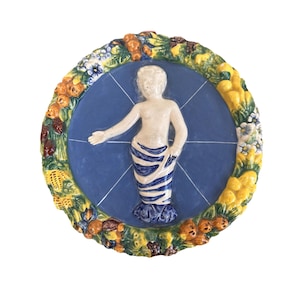 May include: A blue and white ceramic plate featuring a raised design of a figure standing within a circle of flowers and fruit. The figure is wearing a blue and white striped skirt and has a white body.