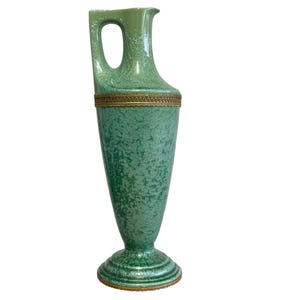 Puede incluir: Un jarrón alto de cerámica verde con asa y pico vertedor. El jarrón tiene una superficie texturizada y una banda dorada en el centro y en la base. El jarrón mide aproximadamente 30 cm de altura.