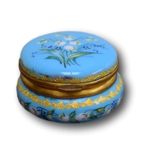May include: A small, round, blue and gold trinket box with a floral design. The lid is decorated with a bouquet of white flowers with green leaves on a light blue background. The box has a gold rim and a gold band around the middle.