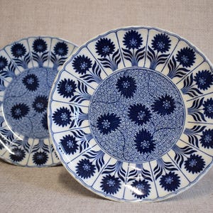 May include: Two white and blue ceramic plates. The plates feature a floral design with dark blue flowers and a detailed pattern in the center. The rims have a scalloped edge. The plates are set against a neutral background.