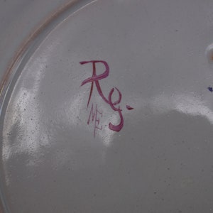 May include: Close-up of a white ceramic plate with a red handwritten signature that reads "Rg." and "ME" in smaller letters.
