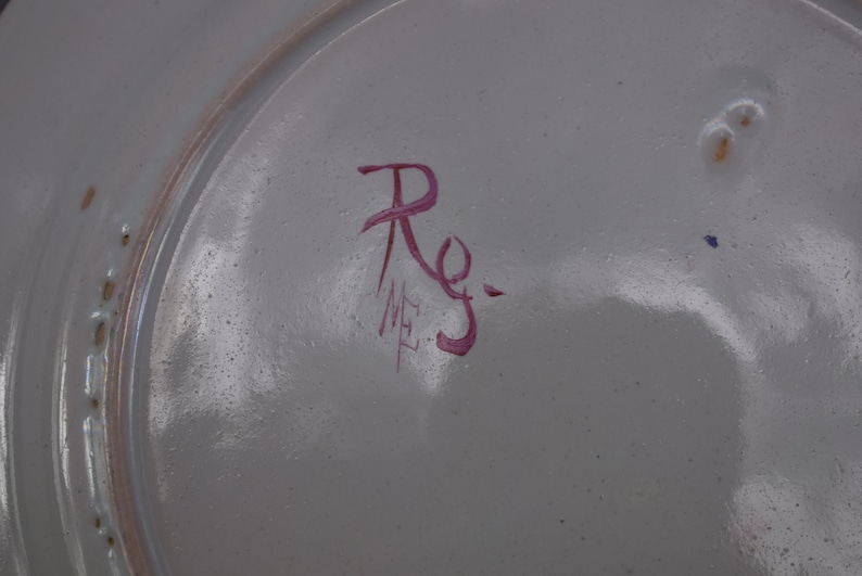 May include: Close-up of a white ceramic plate with a red handwritten signature that reads "Rg." and "ME" in smaller letters.