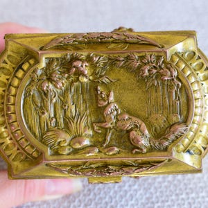 May include: Ornate, rectangular, antique brass box with a detailed relief of a fox in a forest scene. The box has a hinged lid and decorative oval frame. The color is a mix of gold and bronze.