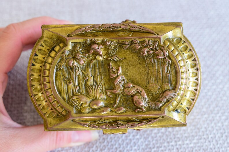 May include: Ornate, rectangular, antique brass box with a detailed relief of a fox in a forest scene. The box has a hinged lid and decorative oval frame. The color is a mix of gold and bronze.