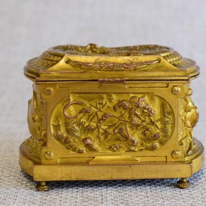 May include: A gold-colored, rectangular box with ornate relief carvings. The box features a hinged lid and rests on small feet. The front panel displays a detailed scene of a lion and foliage. The overall design suggests a vintage or antique style.