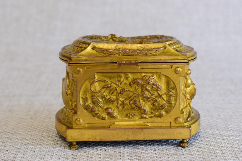 May include: A gold-colored, rectangular box with ornate relief carvings. The box features a hinged lid and rests on small feet. The front panel displays a detailed scene of a lion and foliage. The overall design suggests a vintage or antique style.