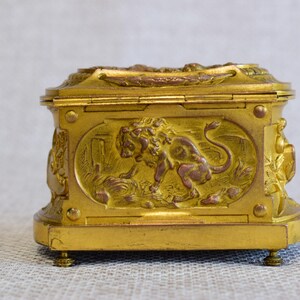 May include: A gold-colored, rectangular box with ornate relief carvings. The box features a lion motif on the front panel and decorative elements on the sides and top. It rests on small, rounded feet, and the overall design suggests a vintage or antique style.
