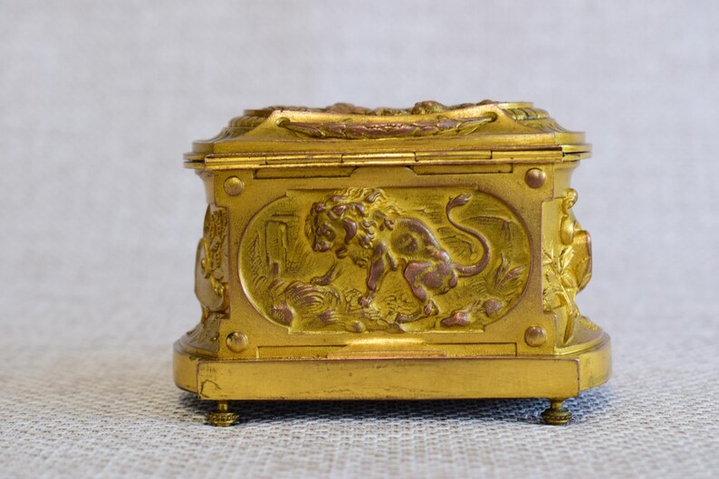 May include: A gold-colored, rectangular box with ornate relief carvings. The box features a lion motif on the front panel and decorative elements on the sides and top. It rests on small, rounded feet, and the overall design suggests a vintage or antique style.