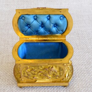 May include: A small, open, gold-colored jewelry box with a blue velvet interior. The box has an oval shape with a hinged lid and is decorated with floral patterns. The interior is lined with a tufted blue velvet fabric.