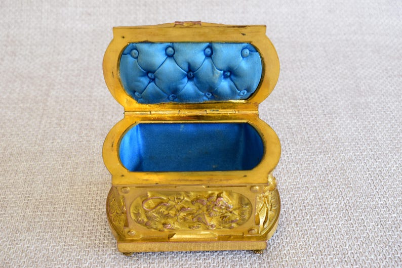 May include: A small, open, gold-colored jewelry box with a blue velvet interior. The box has an oval shape with a hinged lid and is decorated with floral patterns. The interior is lined with a tufted blue velvet fabric.
