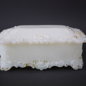 Puede incluir: Una caja de porcelana blanca para alhajas con intrincados detalles dorados. La caja tiene forma cuadrada con una tapa elevada y un borde decorativo festoneado.