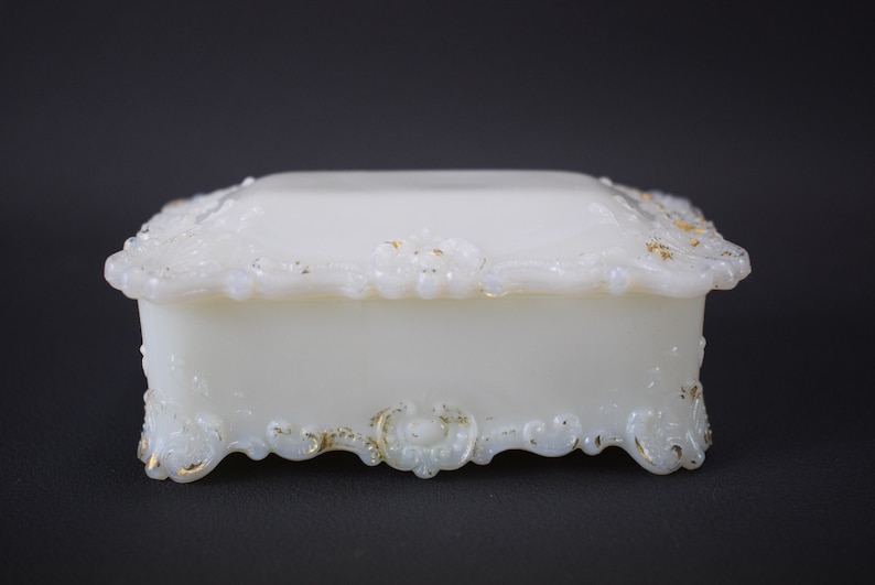 Puede incluir: Una caja de porcelana blanca para alhajas con intrincados detalles dorados. La caja tiene forma cuadrada con una tapa elevada y un borde decorativo festoneado.