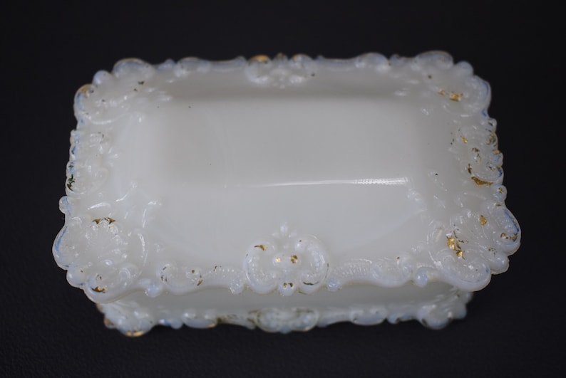 Puede incluir: Caja de porcelana blanca para alhajas con un dise&ntilde;o floral ornamentado y en relieve alrededor de los bordes. La caja tiene un borde dorado.