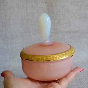 May include: A pink glass powder jar with a white handle on the lid. The jar has a gold-colored band around the rim. The jar is held in a hand.