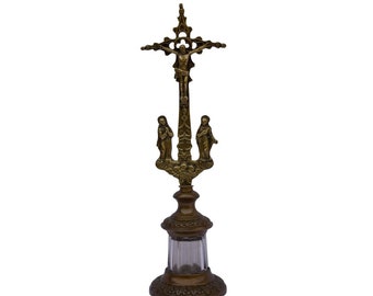 May include: A gold-toned metal crucifix with a figure of Jesus Christ on the cross. The crucifix is mounted on a decorative stand with a clear glass container. The stand has a detailed base with intricate designs.
