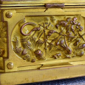 May include: A gold-colored antique box with ornate relief carvings. The box features a detailed lion and floral design on its side, with a hinged lid and decorative feet. The metal box has a tarnished, aged appearance, suggesting it is vintage.