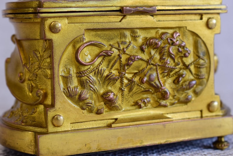 May include: A gold-colored antique box with ornate relief carvings. The box features a detailed lion and floral design on its side, with a hinged lid and decorative feet. The metal box has a tarnished, aged appearance, suggesting it is vintage.