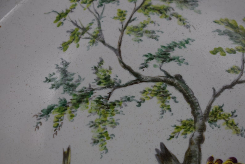 May include: A close-up of a white ceramic plate featuring a hand-painted design of a tree with green leaves. The tree is painted in a realistic style and the leaves are detailed.