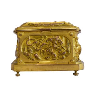 May include: A gold-colored, rectangular box with ornate relief carvings. The box features detailed scenes of figures and animals, with a hinged lid and small feet. The overall design suggests a vintage or antique style, with a focus on decorative elements.