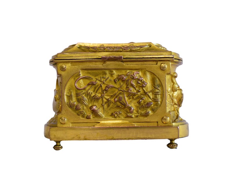 May include: A gold-colored, rectangular box with ornate relief carvings. The box features detailed scenes of figures and animals, with a hinged lid and small feet. The overall design suggests a vintage or antique style, with a focus on decorative elements.