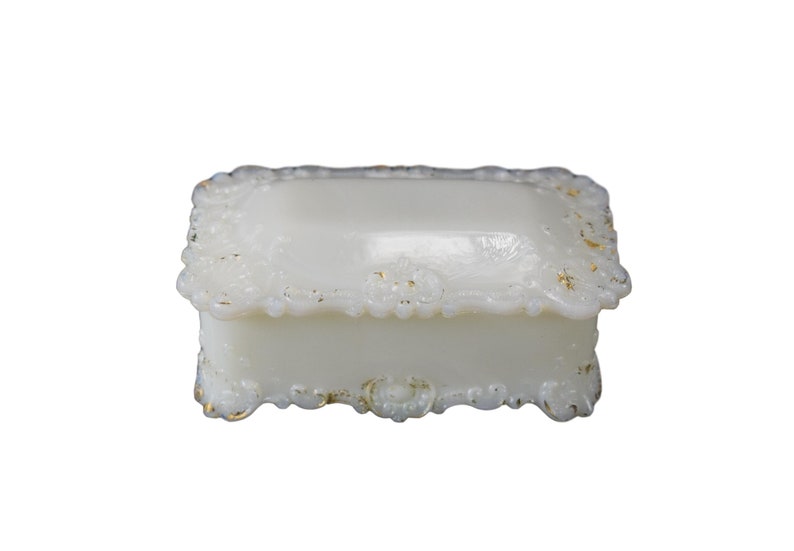 Puede incluir: Una caja de recuerdos de vidrio de leche blanco con un borde dorado y un dise&ntilde;o ornamentado. La caja tiene una forma rectangular y una tapa con bisagras.