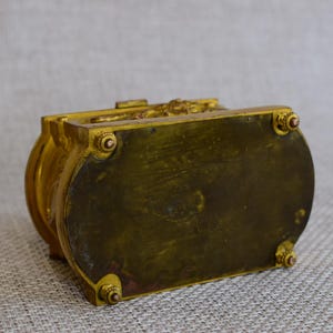 May include: A vintage, ornate box with a gold finish. The box has a rectangular shape with rounded sides and decorative detailing on the top. The bottom of the box has four small feet. The box appears to be made of metal.