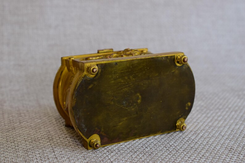 May include: A vintage, ornate box with a gold finish. The box has a rectangular shape with rounded sides and decorative detailing on the top. The bottom of the box has four small feet. The box appears to be made of metal.