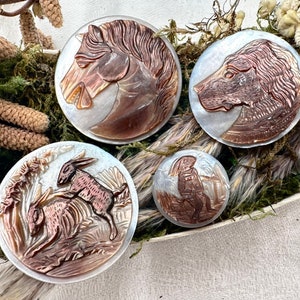 Puede incluir: Cuatro broches redondos de camafeo de concha decorativos con diferentes diseños. Uno presenta una cabeza de caballo, otro una cabeza de perro, otro dos conejos y el último una persona con un sombrero.