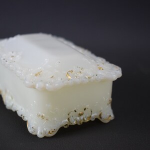 Puede incluir: Una caja de porcelana blanca para alhajas con detalles dorados y dise&ntilde;os florales intrincados. La caja tiene una forma rectangular con una tapa con bisagras y patas ornamentadas.