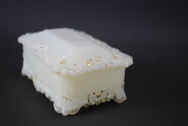 Puede incluir: Una caja de porcelana blanca para alhajas con detalles dorados y dise&ntilde;os florales intrincados. La caja tiene una forma rectangular con una tapa con bisagras y patas ornamentadas.