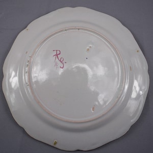 May include: The reverse of a white ceramic plate with a scalloped edge. The plate has a pink and red mark that says "Rg." in cursive.