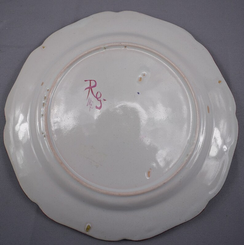 May include: The reverse of a white ceramic plate with a scalloped edge. The plate has a pink and red mark that says "Rg." in cursive.