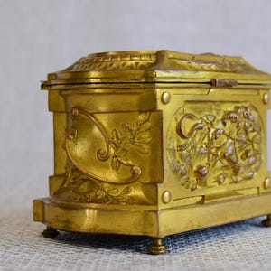 May include: A gold-colored, antique box with ornate relief carvings. The box has a rectangular shape with a hinged lid and rests on small, rounded feet. The sides feature detailed floral and figural designs, adding to its vintage appeal.