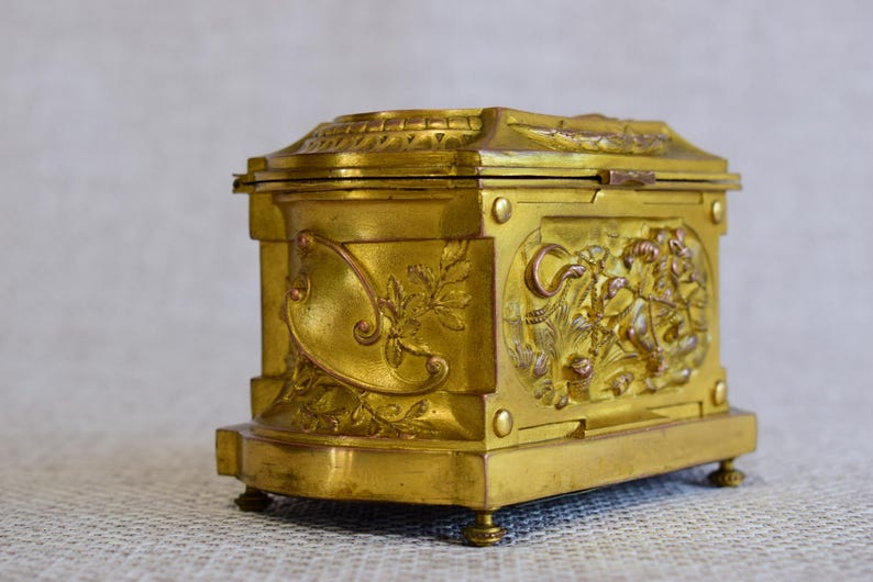 May include: A gold-colored, antique box with ornate relief carvings. The box has a rectangular shape with a hinged lid and rests on small, rounded feet. The sides feature detailed floral and figural designs, adding to its vintage appeal.