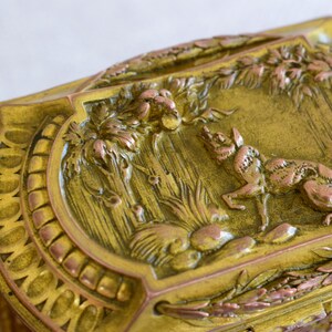 May include: Ornate, antique brass box with a detailed relief scene of a fox in a forest setting. The box features a decorative border and a hinged lid. The relief is in shades of gold and copper, creating a vintage aesthetic.