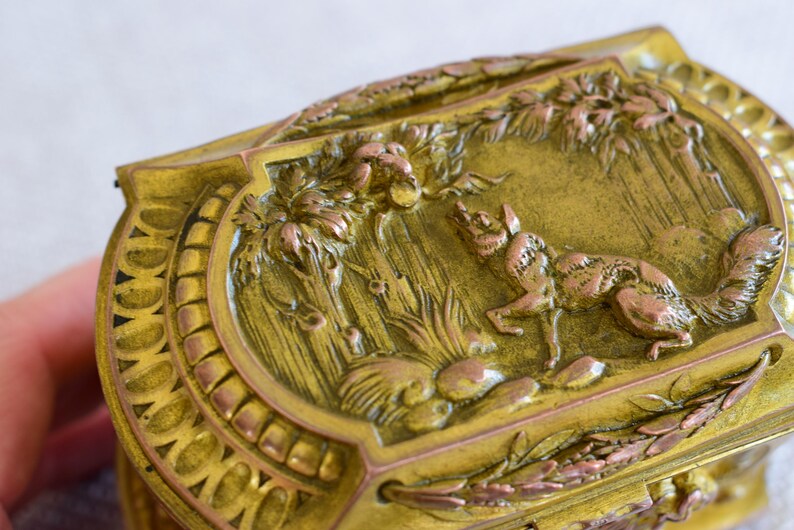 May include: Ornate, antique brass box with a detailed relief scene of a fox in a forest setting. The box features a decorative border and a hinged lid. The relief is in shades of gold and copper, creating a vintage aesthetic.