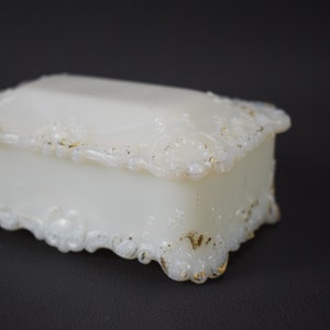 Puede incluir: Una caja de porcelana blanca con ribete dorado y dise&ntilde;os florales intrincados. La caja tiene una tapa con bisagras y se asienta sobre cuatro peque&ntilde;os pies.