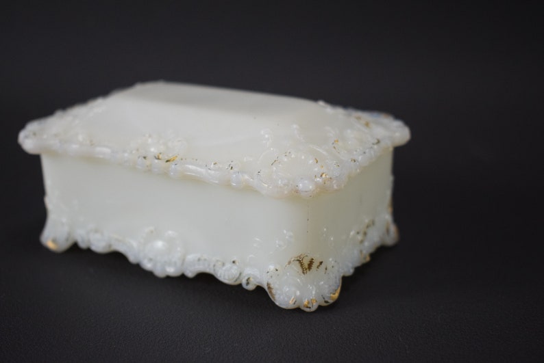 Puede incluir: Una caja de porcelana blanca con ribete dorado y dise&ntilde;os florales intrincados. La caja tiene una tapa con bisagras y se asienta sobre cuatro peque&ntilde;os pies.