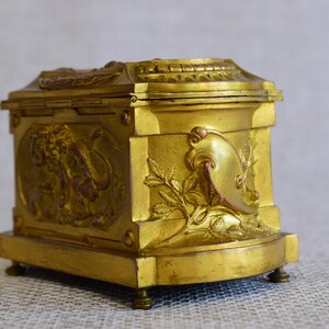 May include: A small, antique gold-colored box with ornate relief details. The box features a hinged lid and is decorated with classical motifs, including a lion and floral elements. It rests on small, rounded feet.