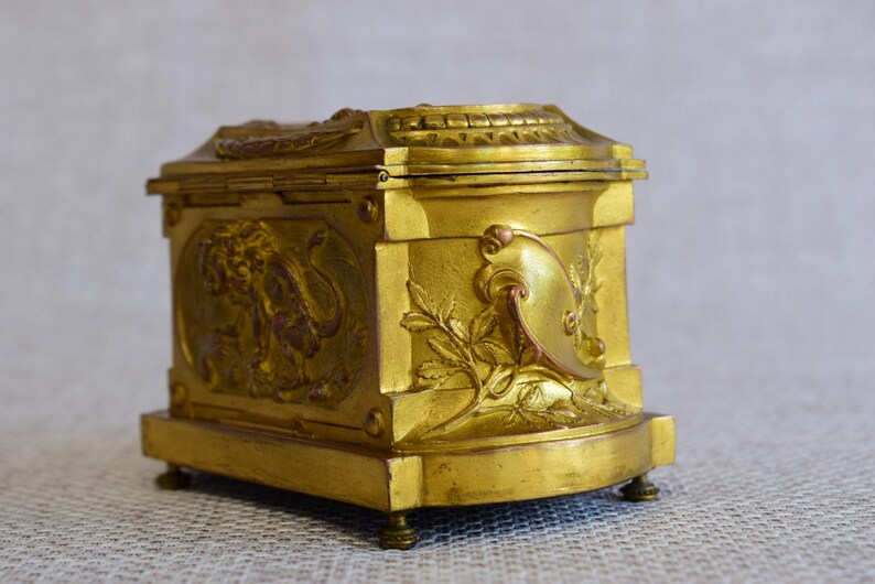 May include: A small, antique gold-colored box with ornate relief details. The box features a hinged lid and is decorated with classical motifs, including a lion and floral elements. It rests on small, rounded feet.