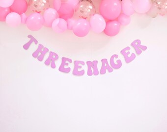 May include: Pink and white balloon garland with a pink banner that reads "THREEnager".