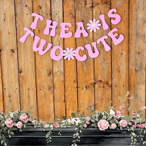 Pu&ograve; includere: Striscione rosa con il testo "Thea is Two Cute" con fiori bianchi tra le parole. Lo striscione &egrave; appeso a una parete di legno con una ghirlanda di fiori sotto.